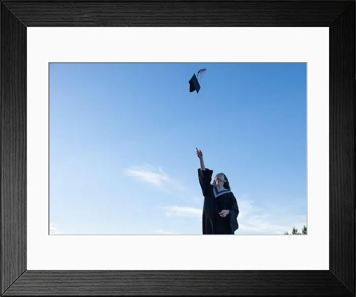Framed Print : Young woman wearing graduation gown tossing her cap, mortar board, into the air with a blue sky in the background Framed Print : Young woman wearing graduation gown tossing her cap, mortar board, into the air with a blue sky in the background