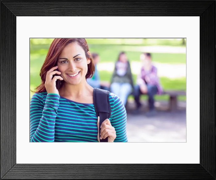 Framed Print : College girl using mobile phone with blurred students in park Framed Print : College girl using mobile phone with blurred students in park