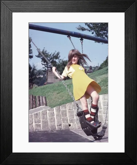 Framed Print : 1970s young girl in yellow dress standing up on a swing in a children's playground Framed Print : 1970s young girl in yellow dress standing up on a swing in a children's playground