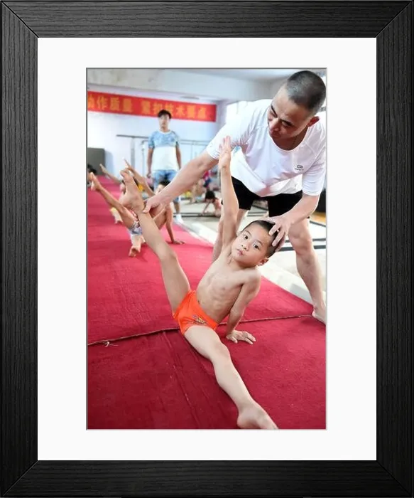 Framed Print : A coach adjusts the posture of a young Chinese boy on the ground to practice gymnastics at a gymnastics training center in Bozhou city, east Chinas An Framed Print : A coach adjusts the posture of a young Chinese boy on the ground to practice gymnastics at a gymnastics training center in Bozhou city, east Chinas An