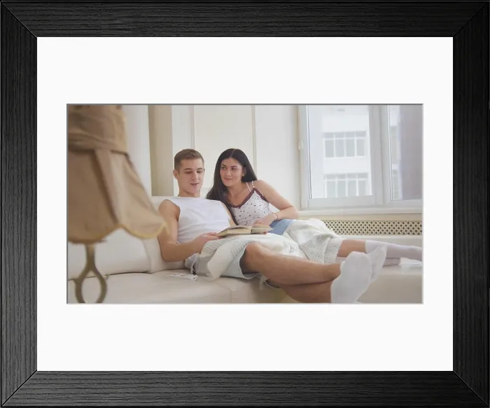 Framed Print : Happy young attractive couple are together on the couch, the girl reads a book, the guy lies on her lap, they're holding hands