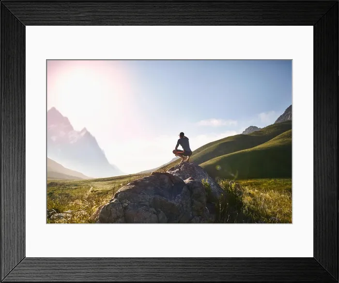 Framed Print : Young man crouching on rock, looking at sunny, r emote mountain view Framed Print : Young man crouching on rock, looking at sunny, r emote mountain view