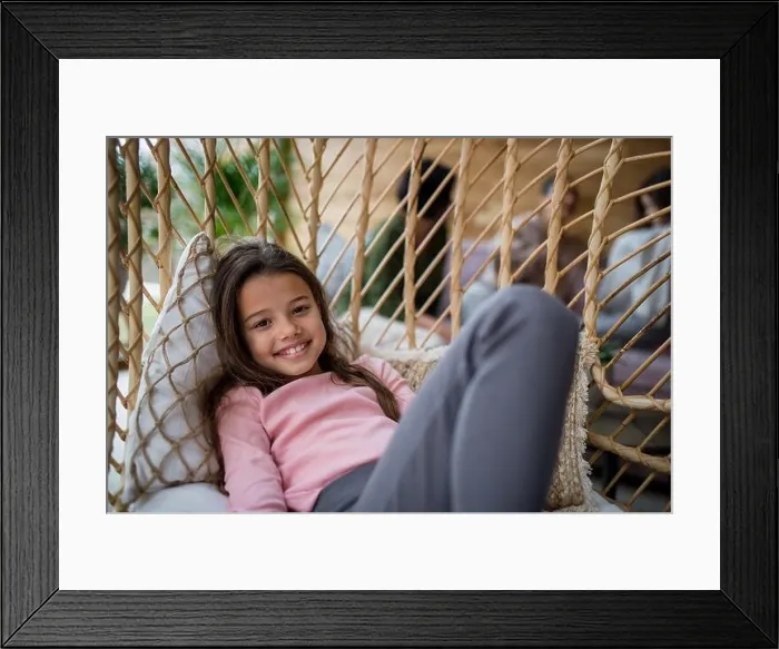 Framed Print : Happy little girl sitting in wicker rattan hang chair outdoors in patio in autumn, looking at camera Framed Print : Happy little girl sitting in wicker rattan hang chair outdoors in patio in autumn, looking at camera