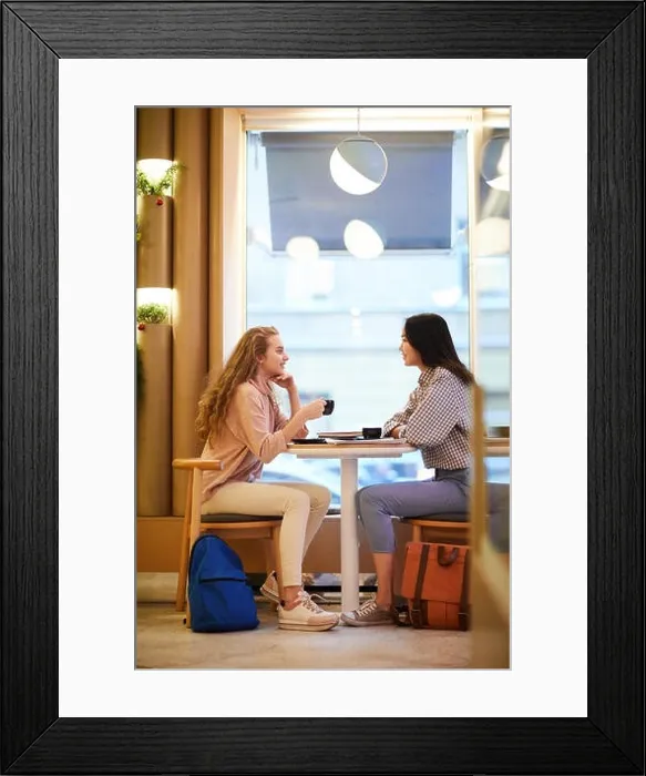 Framed Print : Full length side view portrait of two girls sitting at table in cafe and chatting during break in college, copy space Framed Print : Full length side view portrait of two girls sitting at table in cafe and chatting during break in college, copy space