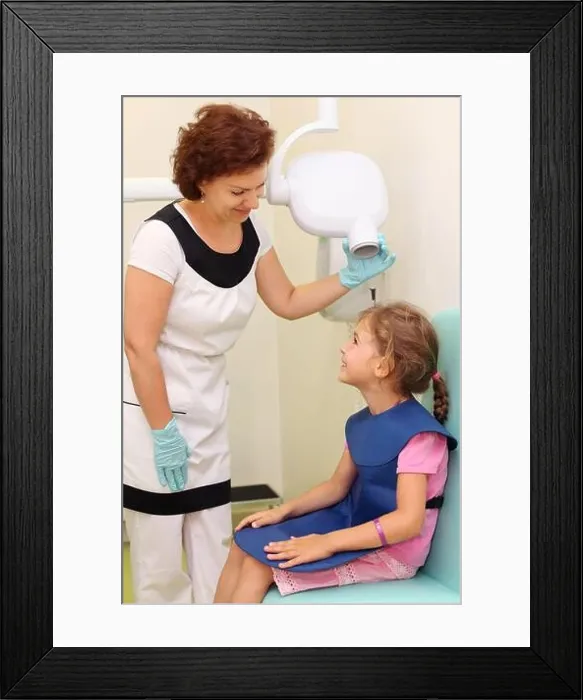 Framed Print : Dentist prepares smiling girl wearing in protective lead apron to jaw x-ray image in dental clinic. Focus on woman Framed Print : Dentist prepares smiling girl wearing in protective lead apron to jaw x-ray image in dental clinic. Focus on woman