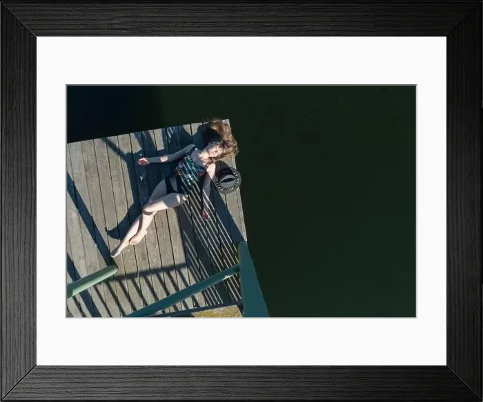 Framed Print : Top view of beautiful Woman in black swimsuit and hat lying down on wooden pier of lake on clear blue sky