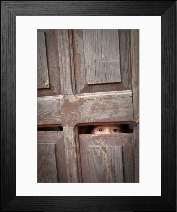 Framed Print : A toddler looks through a small crack in an old wooden door, revealing only her eyes