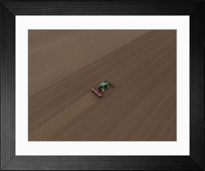 Framed Print : A John Deere Tractor, seen from a distance, cultivates a large field, in readiness for planting Framed Print : A John Deere Tractor, seen from a distance, cultivates a large field, in readiness for planting