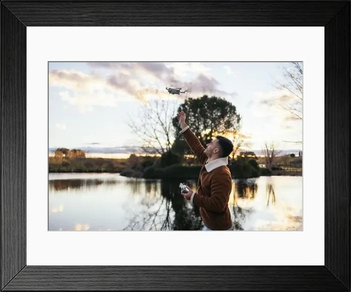 Framed Print : Young man using a drone near a lake to record and photograph the landscape Framed Print : Young man using a drone near a lake to record and photograph the landscape