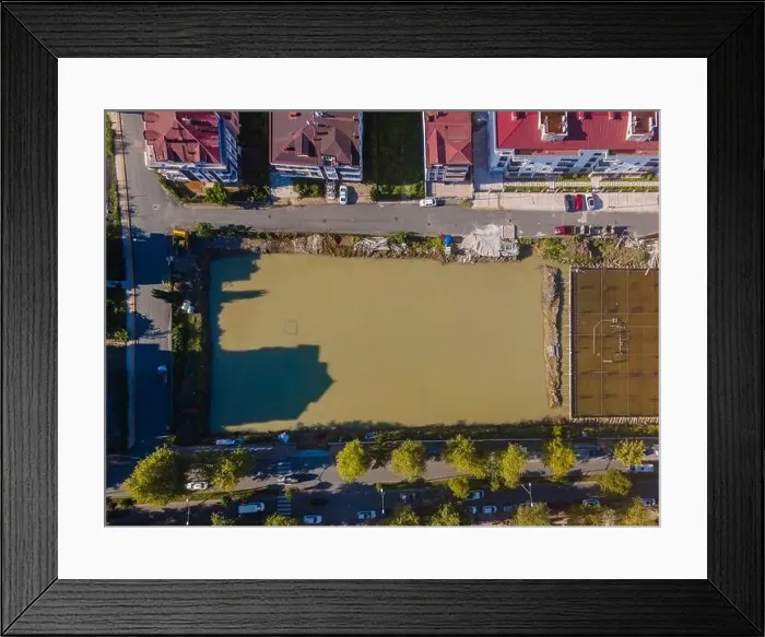 Framed Print : A city street with a large green field in the middle. The field is surrounded by buildings and cars Framed Print : A city street with a large green field in the middle. The field is surrounded by buildings and cars