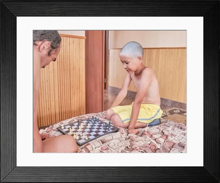 Framed Print : Asian tanned boy with a haircut bald, in yellow shorts, playing chess with an adult on the bed at home Framed Print : Asian tanned boy with a haircut bald, in yellow shorts, playing chess with an adult on the bed at home
