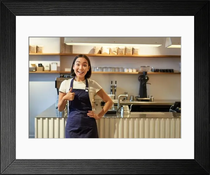 Framed Print : Smiling girl student working part-time in cafe, barista shows thumbs up, wears apron, stands near coffee shop counter Framed Print : Smiling girl student working part-time in cafe, barista shows thumbs up, wears apron, stands near coffee shop counter