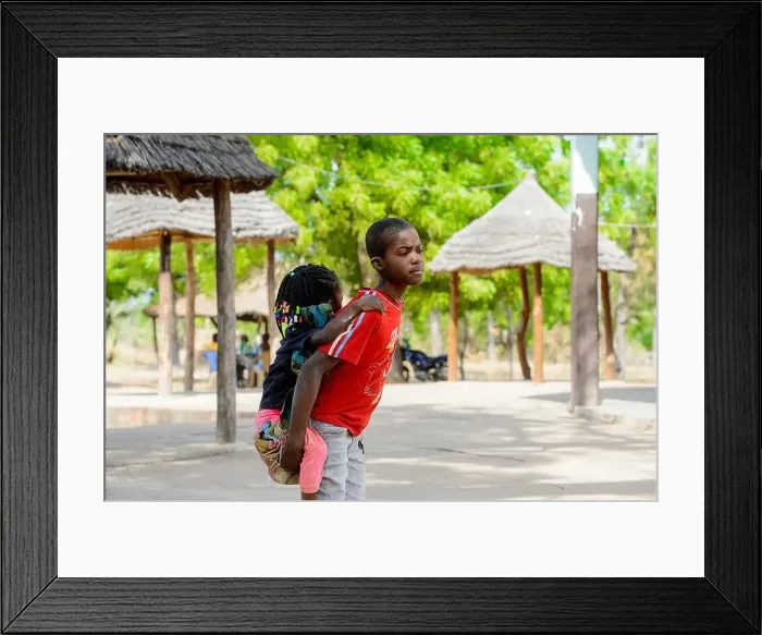 Framed Print : KOUTAMMAKOU, TOGO - JAN 13, 2017: Unidentified Togolese boy in red shirt carries his little sister in the village Framed Print : KOUTAMMAKOU, TOGO - JAN 13, 2017: Unidentified Togolese boy in red shirt carries his little sister in the village
