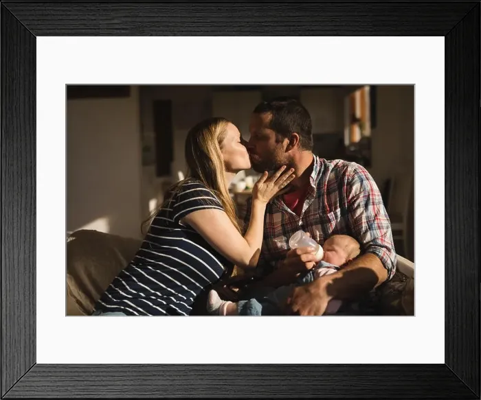Framed Print : Parents kissing each other while father feeding to baby on sofa Framed Print : Parents kissing each other while father feeding to baby on sofa