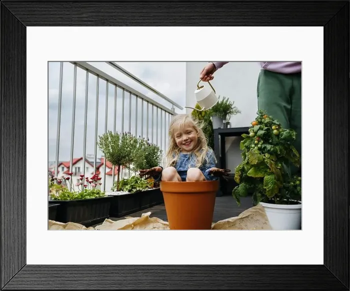 Framed Print : Father watering playful girl sitting in pot at balcony Framed Print : Father watering playful girl sitting in pot at balcony