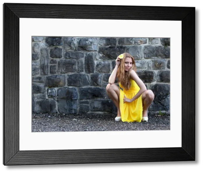Framed Print : Young woman in a yellow dress and a yellow flower posing in a crouching position in front of a stone wall Framed Print : Young woman in a yellow dress and a yellow flower posing in a crouching position in front of a stone wall