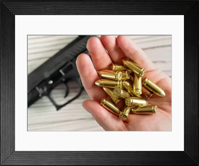Framed Print : Yellow golden coloured bullets held in hand, blurred wooden desk with black gun under Framed Print : Yellow golden coloured bullets held in hand, blurred wooden desk with black gun under