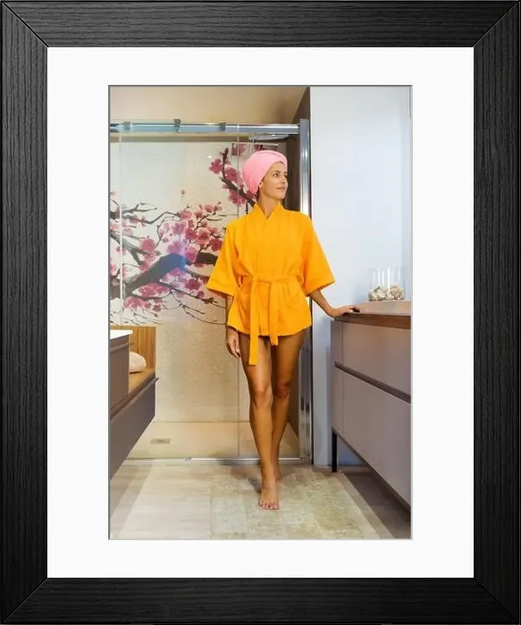 Framed Print : Young woman wearing a colorful orange bathrobe with her hair wrapped in a towel exiting a glass shower cubicle looking