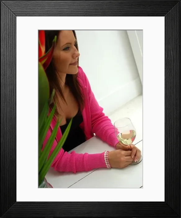 Framed Print : Young woman sat down clasping a glass of white wine on a white table top Framed Print : Young woman sat down clasping a glass of white wine on a white table top