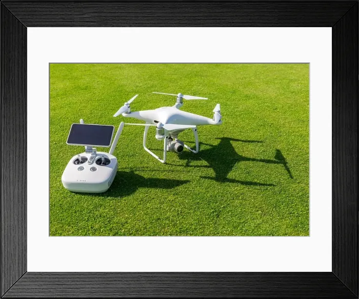 Framed Print : White professional quadcopter drone camera with remote control from above against green grass in background Framed Print : White professional quadcopter drone camera with remote control from above against green grass in background
