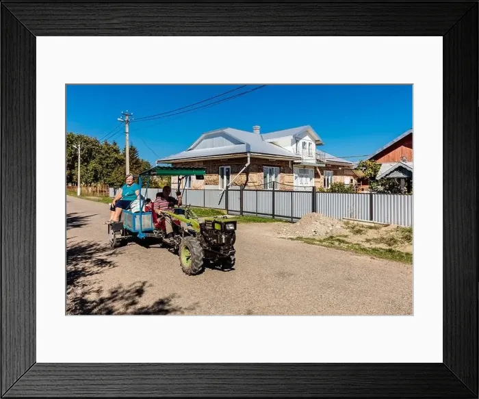 Framed Print : A woman and a man are riding in a cart pulled by a tractor. The cart is on a dirt road in front of a house Framed Print : A woman and a man are riding in a cart pulled by a tractor. The cart is on a dirt road in front of a house