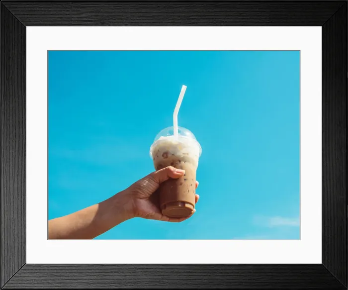 Framed Print : Hand holding a cup of iced coffee on blue sky background Framed Print : Hand holding a cup of iced coffee on blue sky background