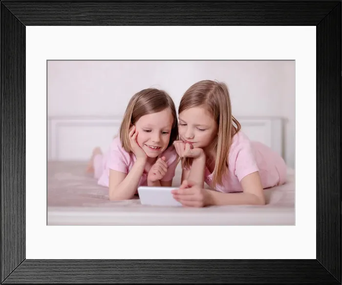 Framed Print : Two little girls (sisters) in pink pajamas on the bed watching a smartphone and reading an e-book. Selective focus