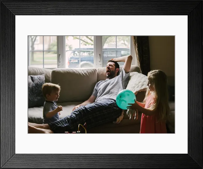 Framed Print : A tired father lies on the couch while his children play around him