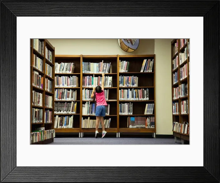 Framed Print : Girl reaching for book on library shelf Framed Print : Girl reaching for book on library shelf