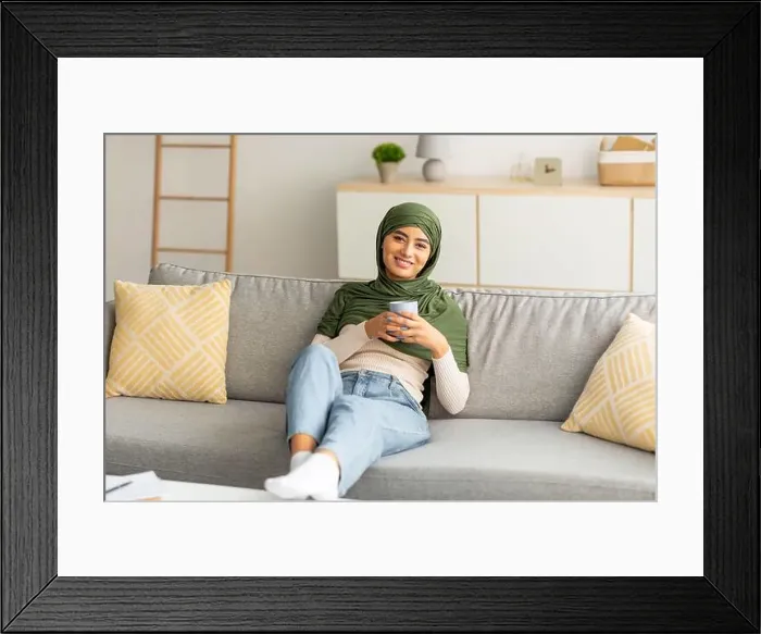 Framed Print : Peaceful weekend. Positive young Arab lady in hijab relaxing on sofa with cozy hot drink at home Framed Print : Peaceful weekend. Positive young Arab lady in hijab relaxing on sofa with cozy hot drink at home