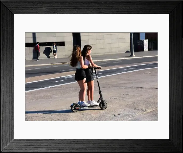 Framed Print : Two teenage girls riding together an electric scooter in a street of a city Framed Print : Two teenage girls riding together an electric scooter in a street of a city