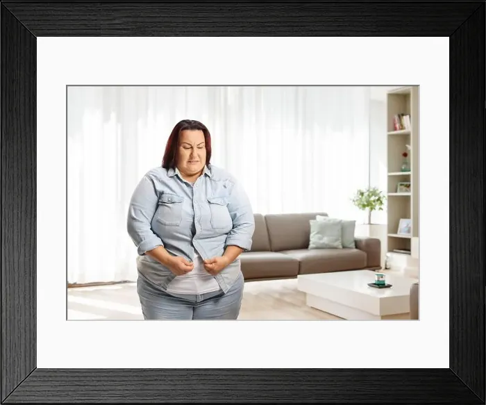 Framed Print : Irritated overweight woman at home trying to button a denim shirt Framed Print : Irritated overweight woman at home trying to button a denim shirt