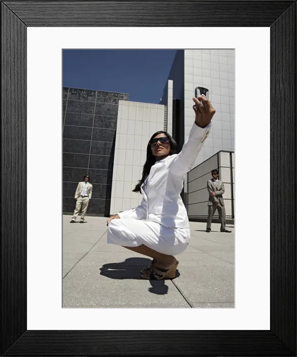 Framed Print : A woman poses with a cellphone while two men are seen in the background Framed Print : A woman poses with a cellphone while two men are seen in the background