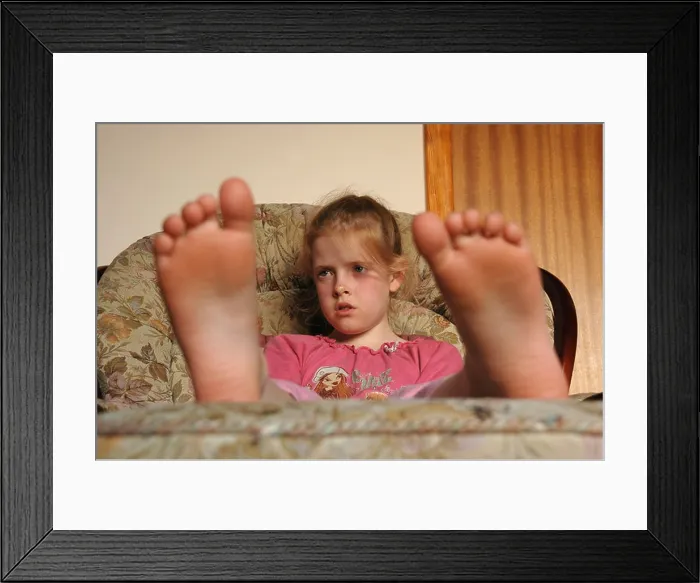 Framed Print : A young seated girl, watches T.V with her bare feet resting on a stool