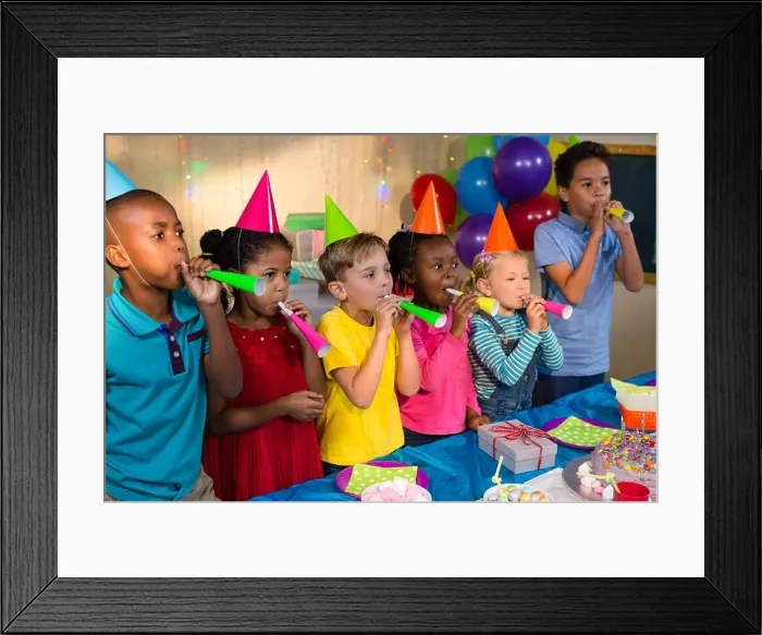 Framed Print : Playful children blowing party horns while standing by table during birthday party Framed Print : Playful children blowing party horns while standing by table during birthday party
