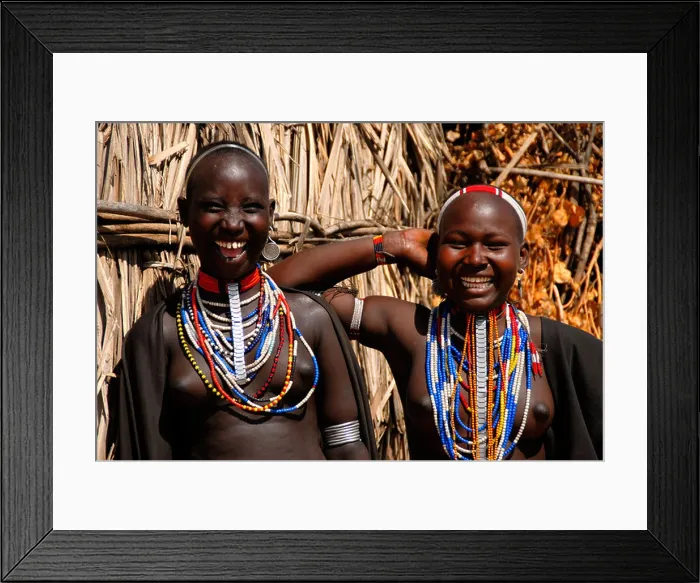 Framed Print : Two young laughing girls of the Arbore people wearing many colourful necklaces in front of a hut Ethiopia