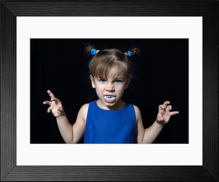 Framed Print : cute little vampire girl in blue dress with big fangs making scary faces at All Hallows Eve party on black background