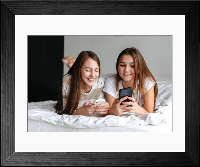Framed Print : Two cute smiling teengae girls laying on bed, holding mobile phones at home Framed Print : Two cute smiling teengae girls laying on bed, holding mobile phones at home