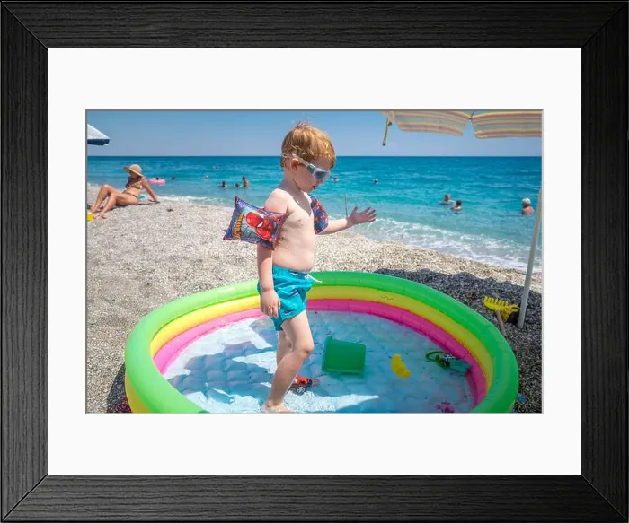 Framed Print : Cute little boy playing in his inflatable pool wearing inflatable armbands and swimming goggles in the beach Framed Print : Cute little boy playing in his inflatable pool wearing inflatable armbands and swimming goggles in the beach