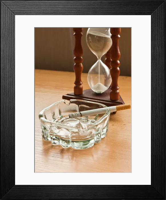 Framed Print : Hourglass and smoking a cigarette in a glass ashtray on wooden top Framed Print : Hourglass and smoking a cigarette in a glass ashtray on wooden top