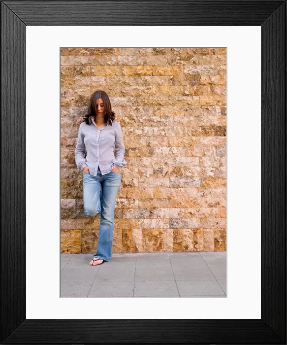 Framed Print : F.S. of young woman with striped shirt, blue jeans and sandals, standing and leaning against a stone wall, looking down