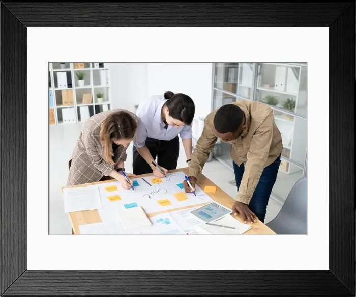 Framed Print : Three intercultural colleagues with highlighters bending over table with papers Framed Print : Three intercultural colleagues with highlighters bending over table with papers
