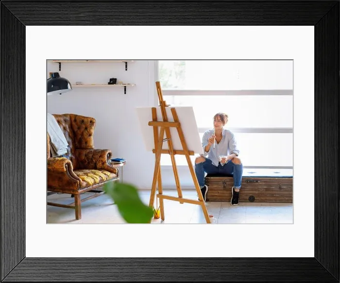 Framed Print : Thoughtful female painter looking at painting while sitting in studio Framed Print : Thoughtful female painter looking at painting while sitting in studio