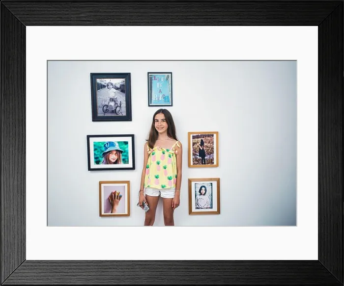 Framed Print : young girl with camera standing against picture frames on wall at home Framed Print : young girl with camera standing against picture frames on wall at home