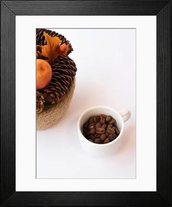 Framed Print : Small white cup of coffee beans next to a fall decoration made of pine cones
