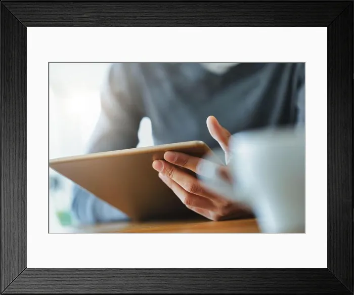 Framed Print : Hand of a Black man holding a tablet pc in close up viewed past a mug of coffee on a table Framed Print : Hand of a Black man holding a tablet pc in close up viewed past a mug of coffee on a table