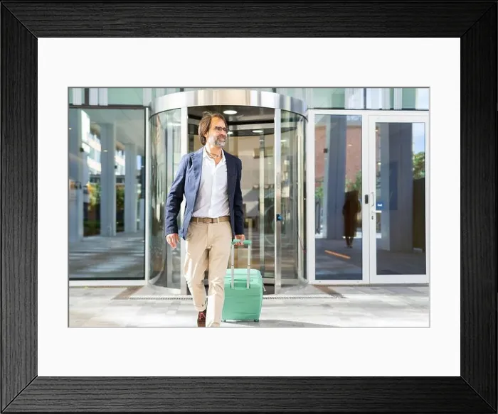 Framed Print : A mature businessman confidently exits a modern glass building, pulling along a light green suitcase and looking away Framed Print : A mature businessman confidently exits a modern glass building, pulling along a light green suitcase and looking away