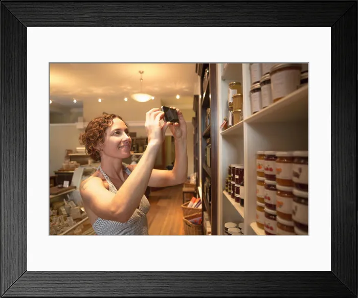 Framed Print : Mid adult woman photographing preserves on smartphone in organic grocery Framed Print : Mid adult woman photographing preserves on smartphone in organic grocery