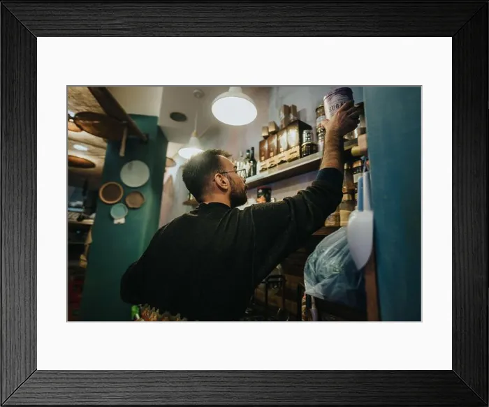 Framed Print : Man reaching for a product on a shelf in a cozy small store Framed Print : Man reaching for a product on a shelf in a cozy small store