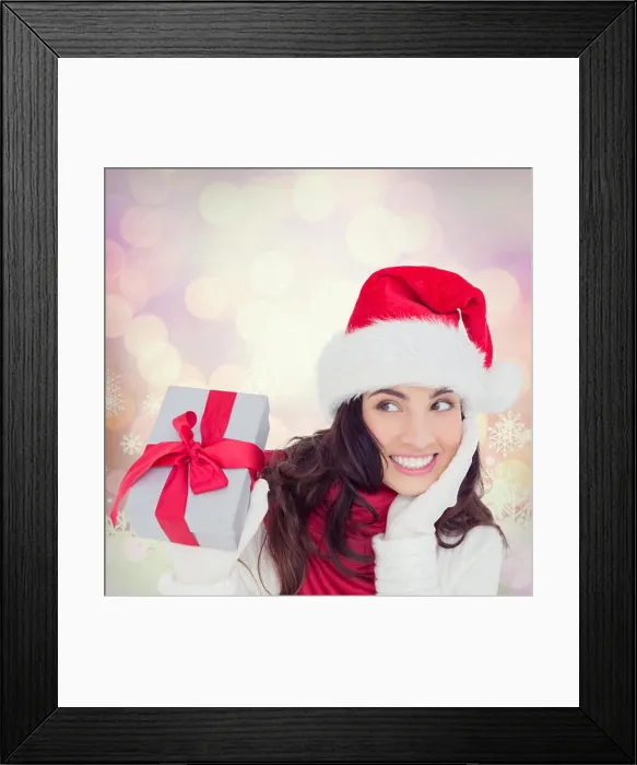 Framed Print : Composite image of surprised brunette in santa hat holding gift Framed Print : Composite image of surprised brunette in santa hat holding gift